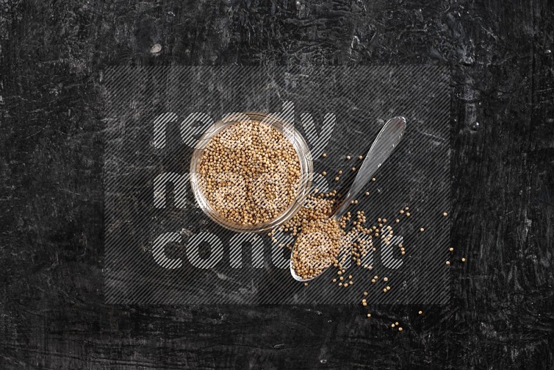 A glass jar and a metal spoon full of mustard seeds on a textured black flooring in different angles