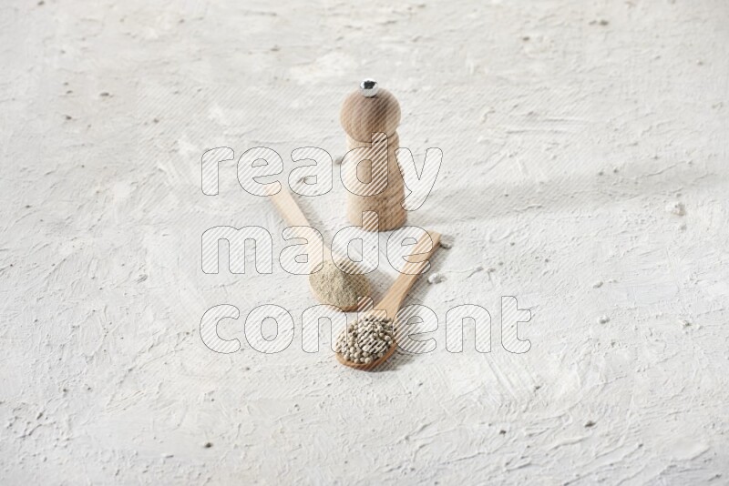 2 wooden spoons one full of white pepper powder and the other with pepper beads and a wooden pepper grinder on textured white flooring