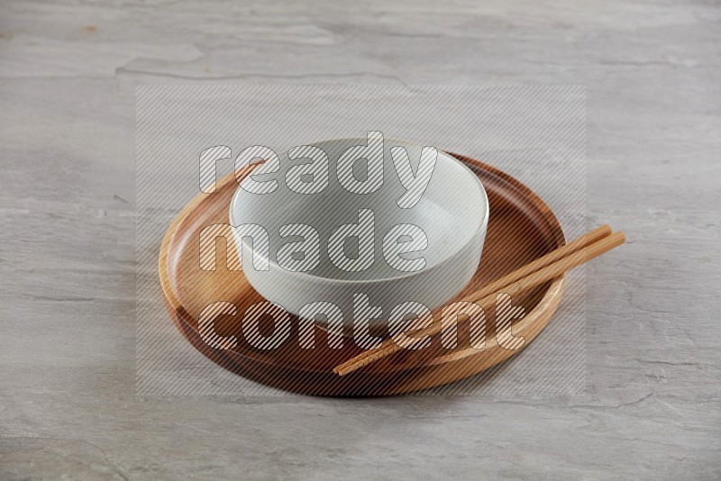 white pottery round bowl on top of brown wood round plate and wood chopsticks, on grey textured countertop