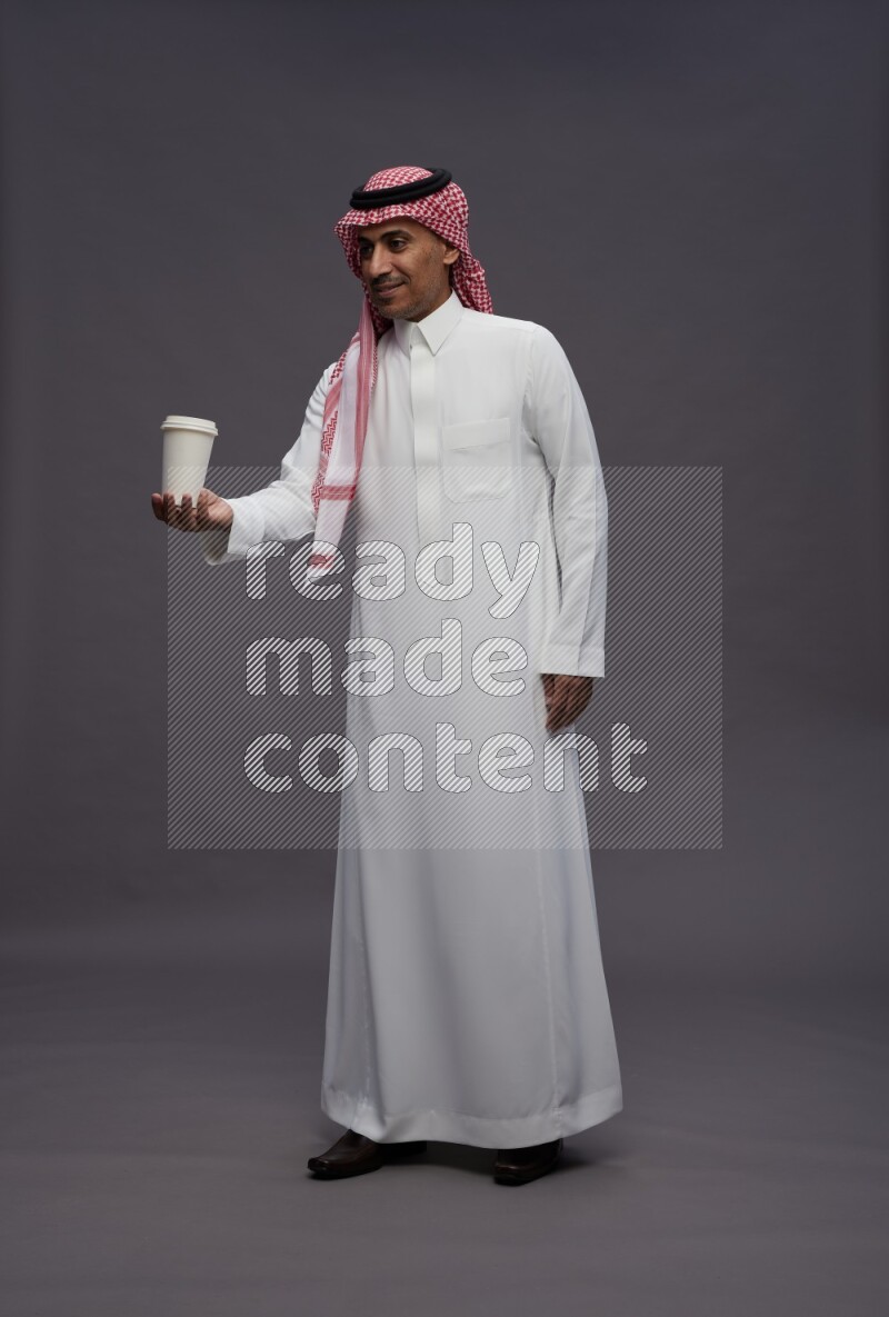 Saudi man wearing thob and shomag standing holding paper cup on gray background