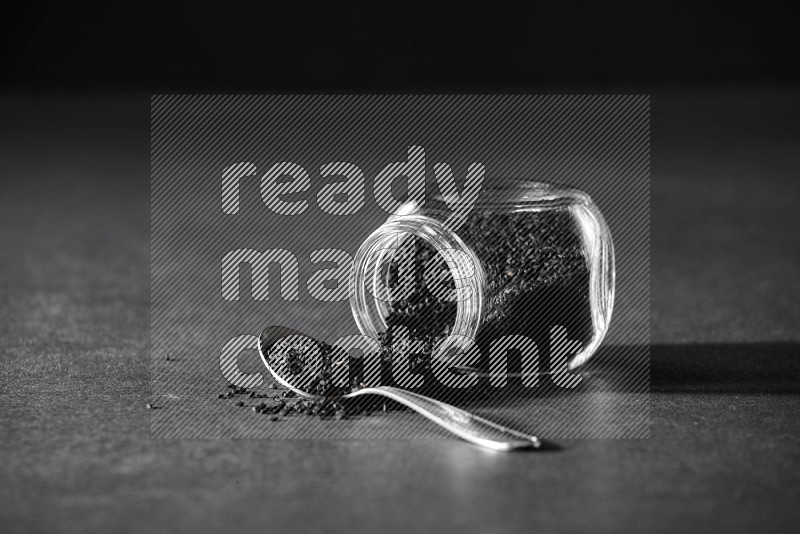 A flipped glass spice jar full of black seeds and the seeds spread out with a metal spoon full it on a black flooring