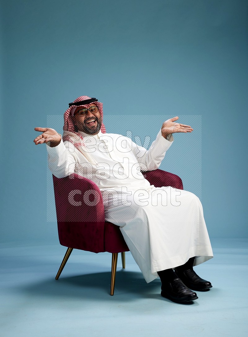 Saudi Man with shimag sitting on chair Interacting with the camera on blue background