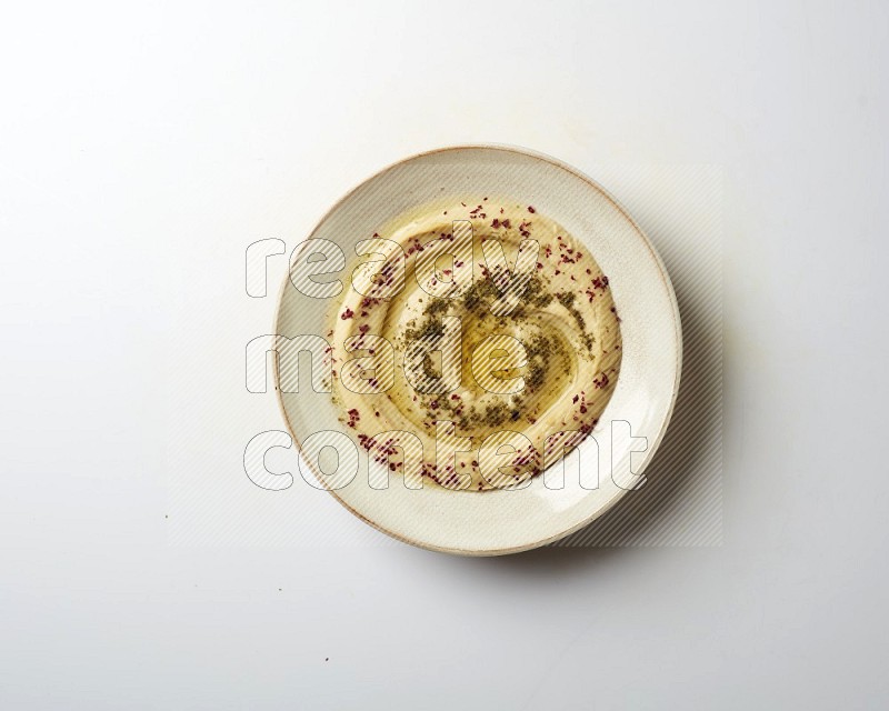 Hummus in a pottry plate garnished with zaatr & sumak on a white background