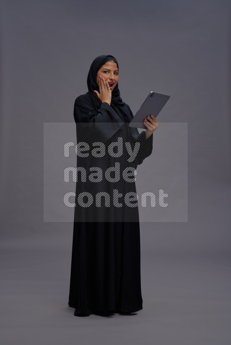 Saudi woman wearing Abaya standing working on tablet on gray background