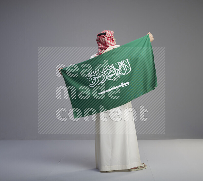 A saudi man standing wearing thob and red shomag holding big saudi flag on gray background