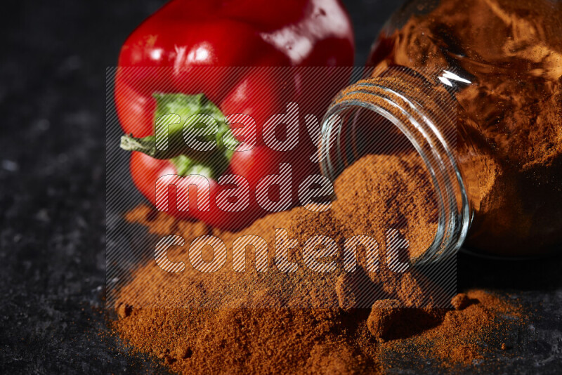 A glass jar full of ground paprika powder flipped with some spilling powder on black background