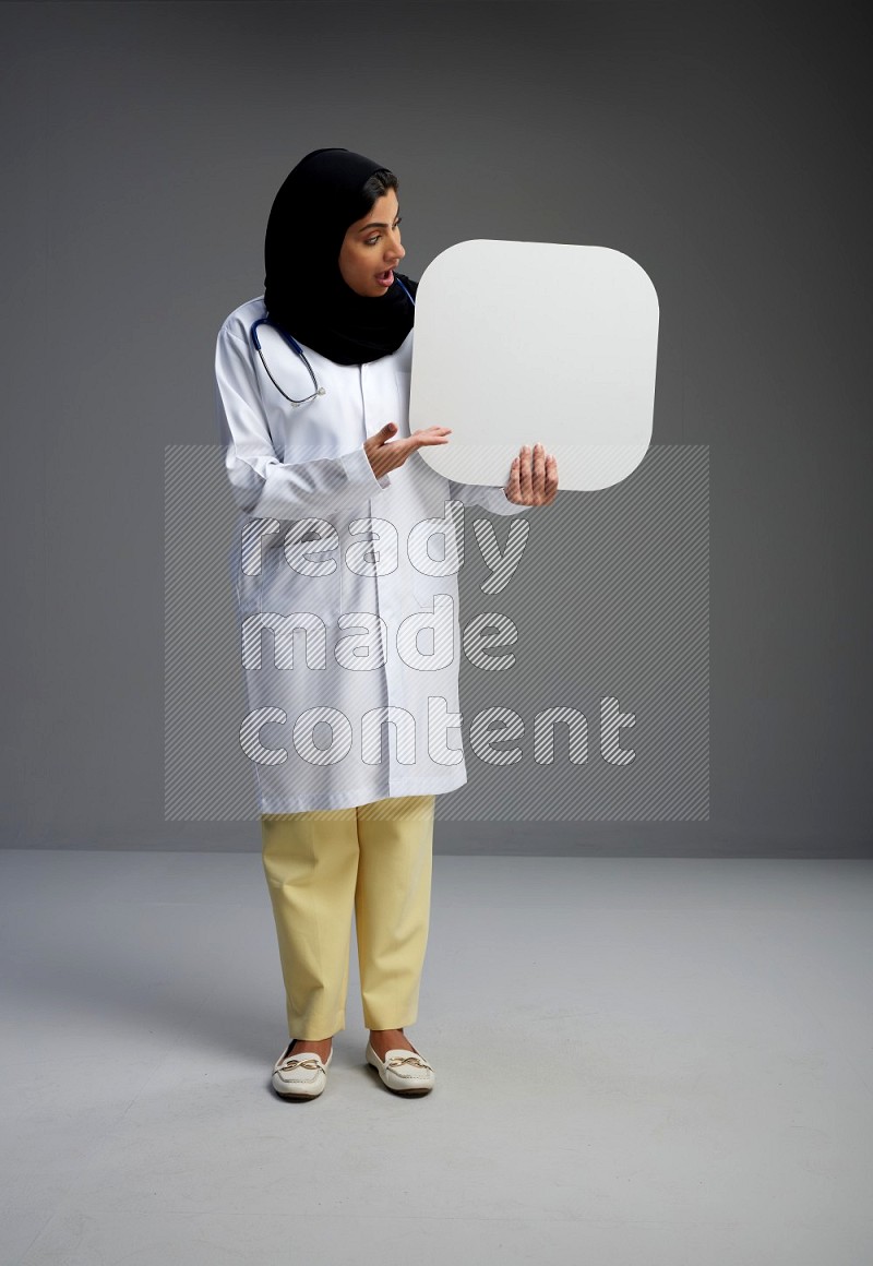 Saudi woman wearing lab coat with stethoscope standing holding social media sign on Gray background