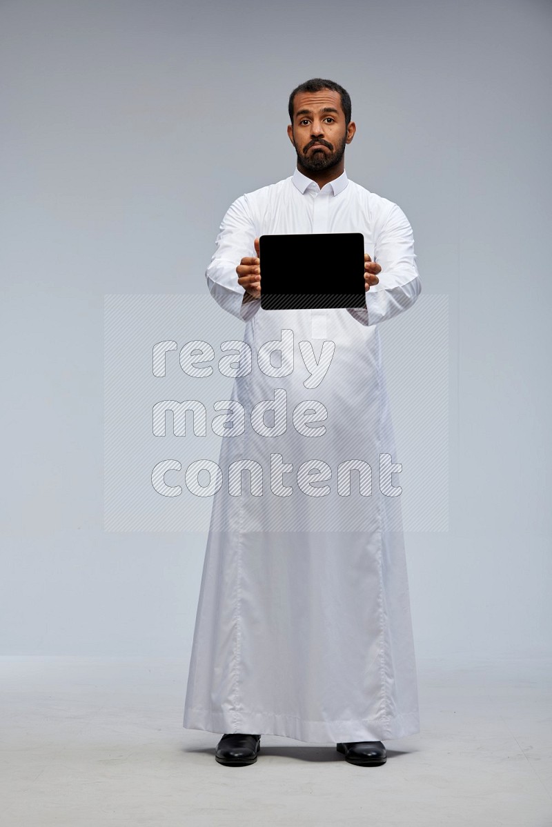 Saudi man wearing Thob standing showing tablet to camera on Gray background