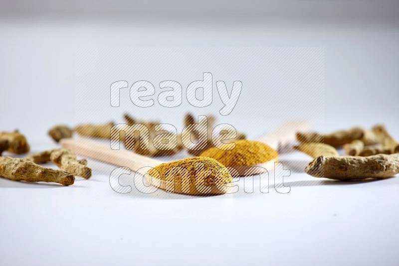 2 wooden spoons full of turmeric powder with dried turmeric fingers beside it on white flooring