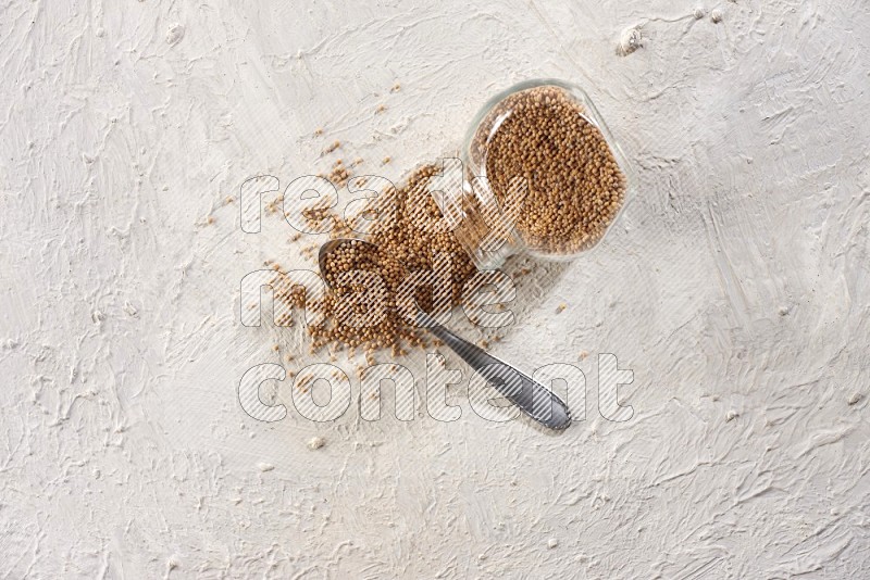 A glass spice jar and a metal spoon full of mustard seeds and jar is flipped with fallen seeds on a textured white flooring
