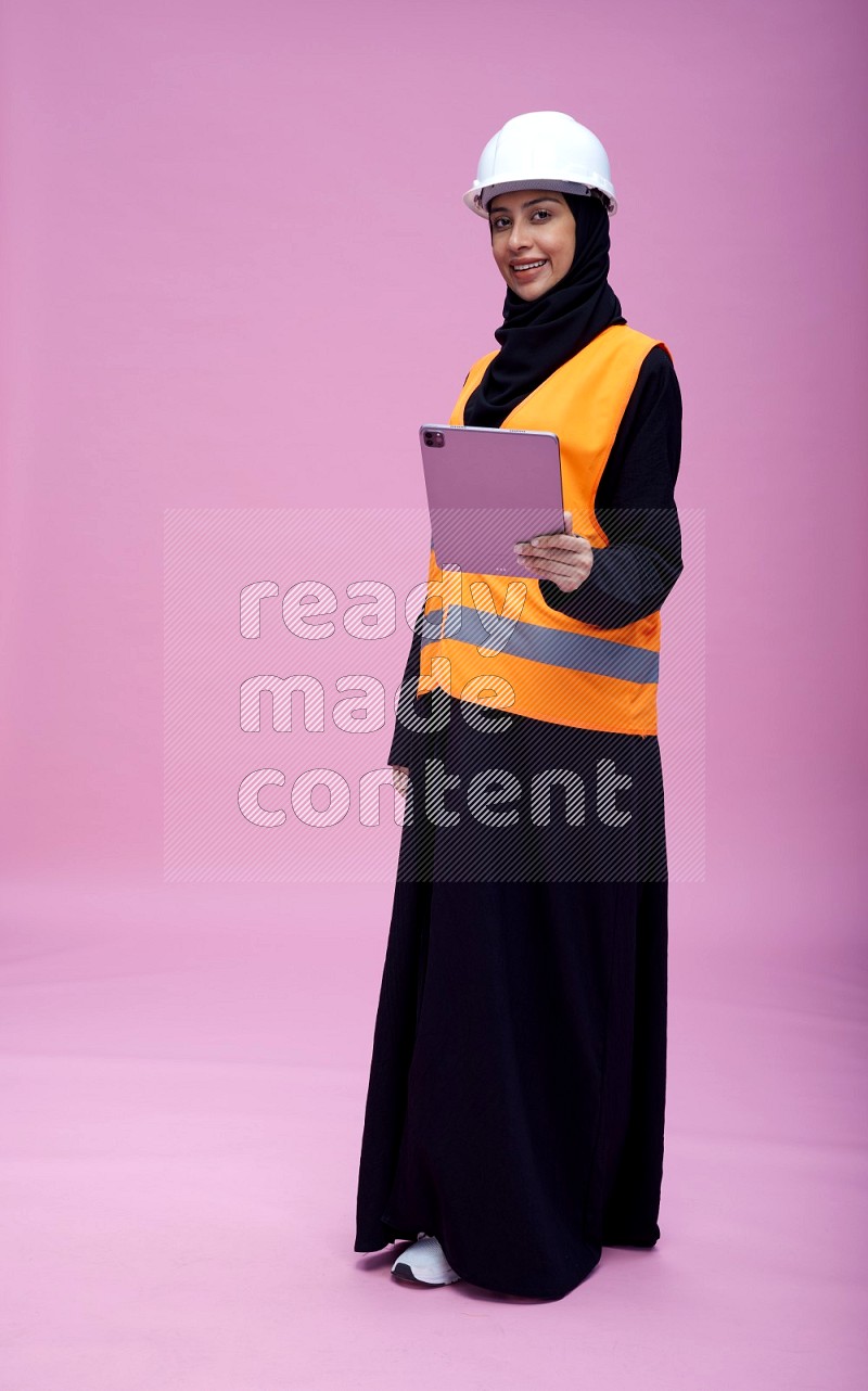Saudi woman wearing Abaya with engineer vest and helmet standing working on tablet on pink background