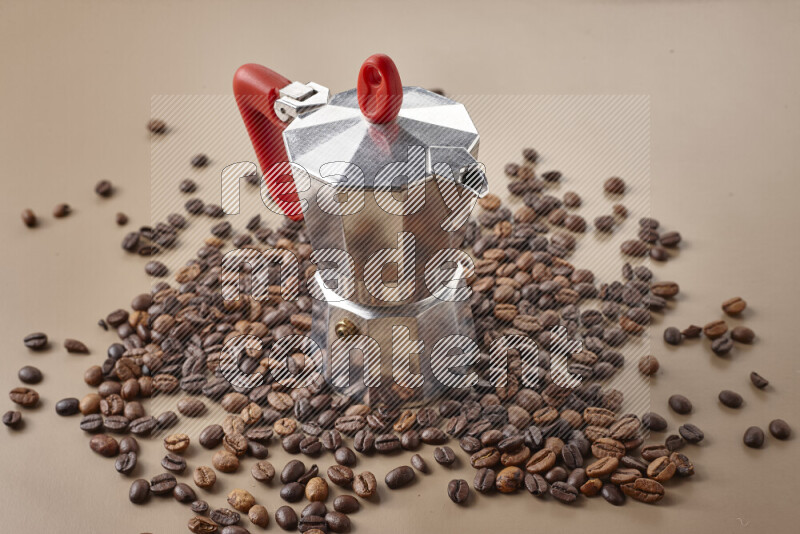 A moka pot with red handle surrounded by roasted coffee beans on beige background