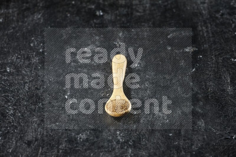 A wooden spoons full of cumin powder on textured black flooring