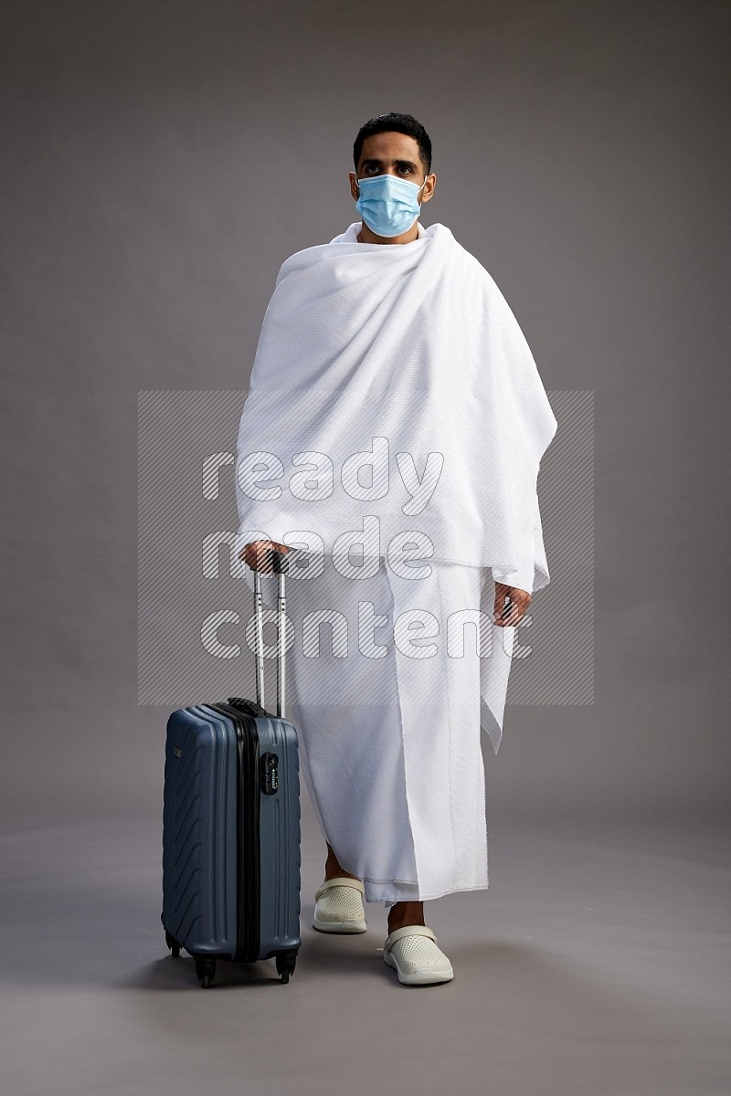A man wearing Ehram with face mask Standing holding traveling bag on gray background
