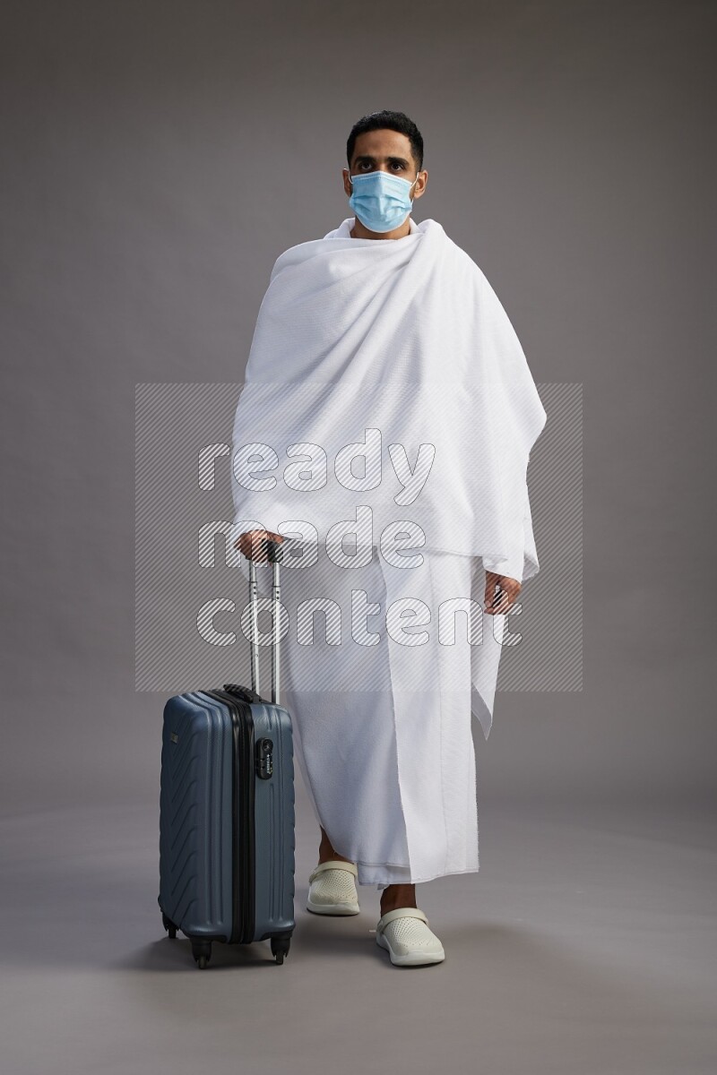 A man wearing Ehram with face mask Standing holding traveling bag on gray background