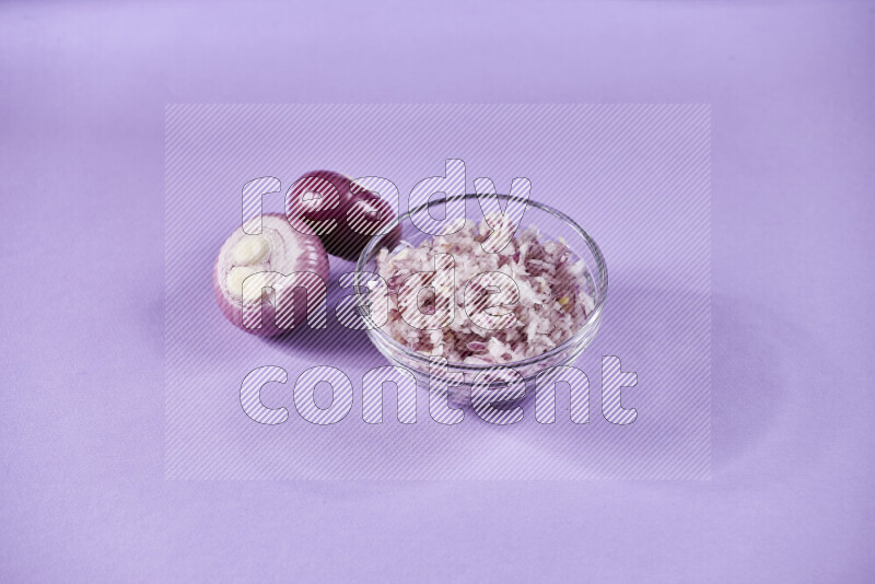 A glass bowl full of chopped red onions with halved onions beside it on purple background