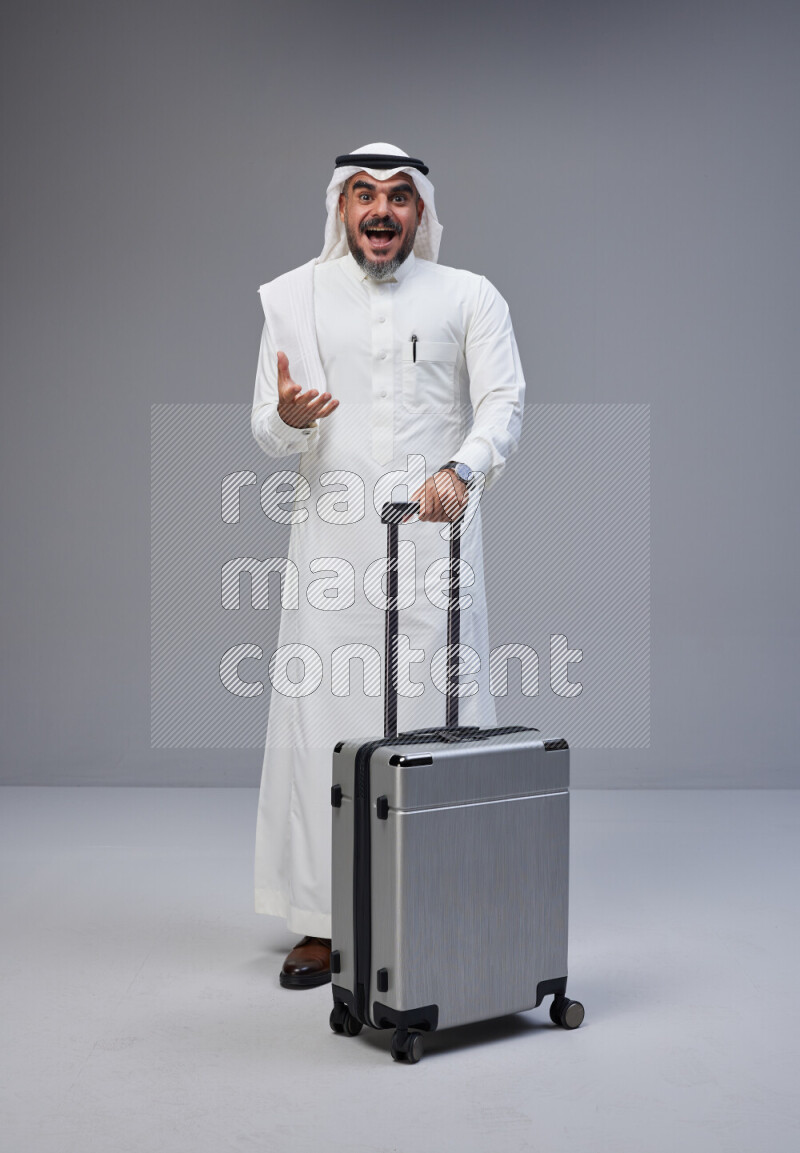 Saudi man wearing Thob and white Shomag standing holding Travel bag on Gray background