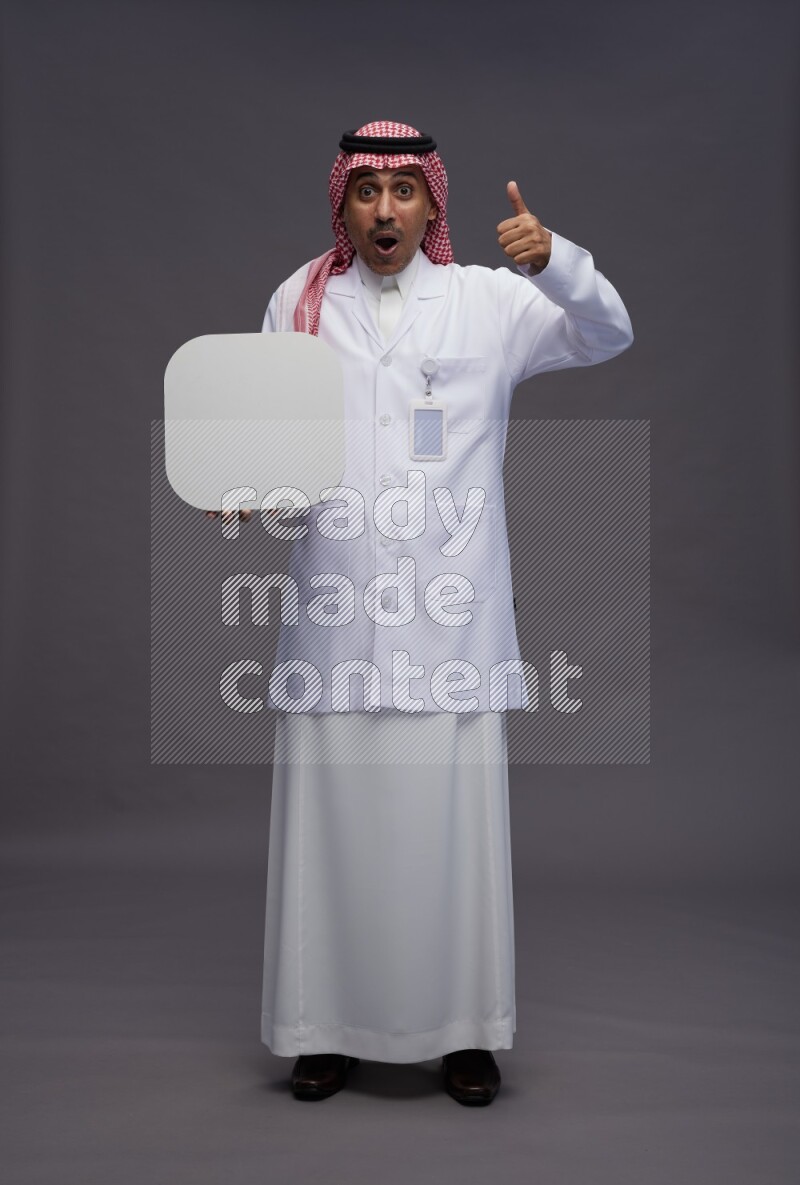 Saudi man wearing thob with lab coat and shomag with pocket employee badge standing holding social media sign on gray background