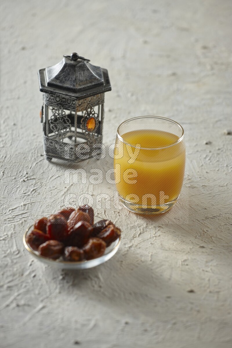 A silver lantern with different drinks, dates, nuts, prayer beads and quran on textured white background