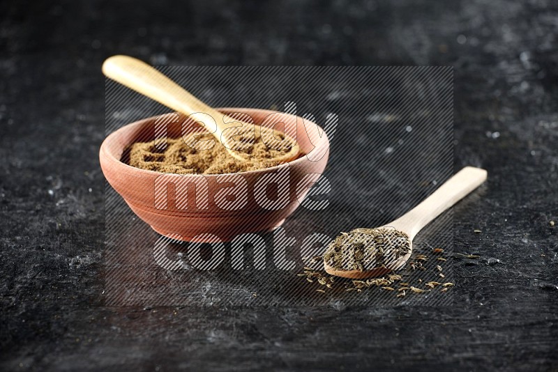 A wooden bowl and 2 wooden spoons full of cumin powder and cumin seeds on a textured black flooring