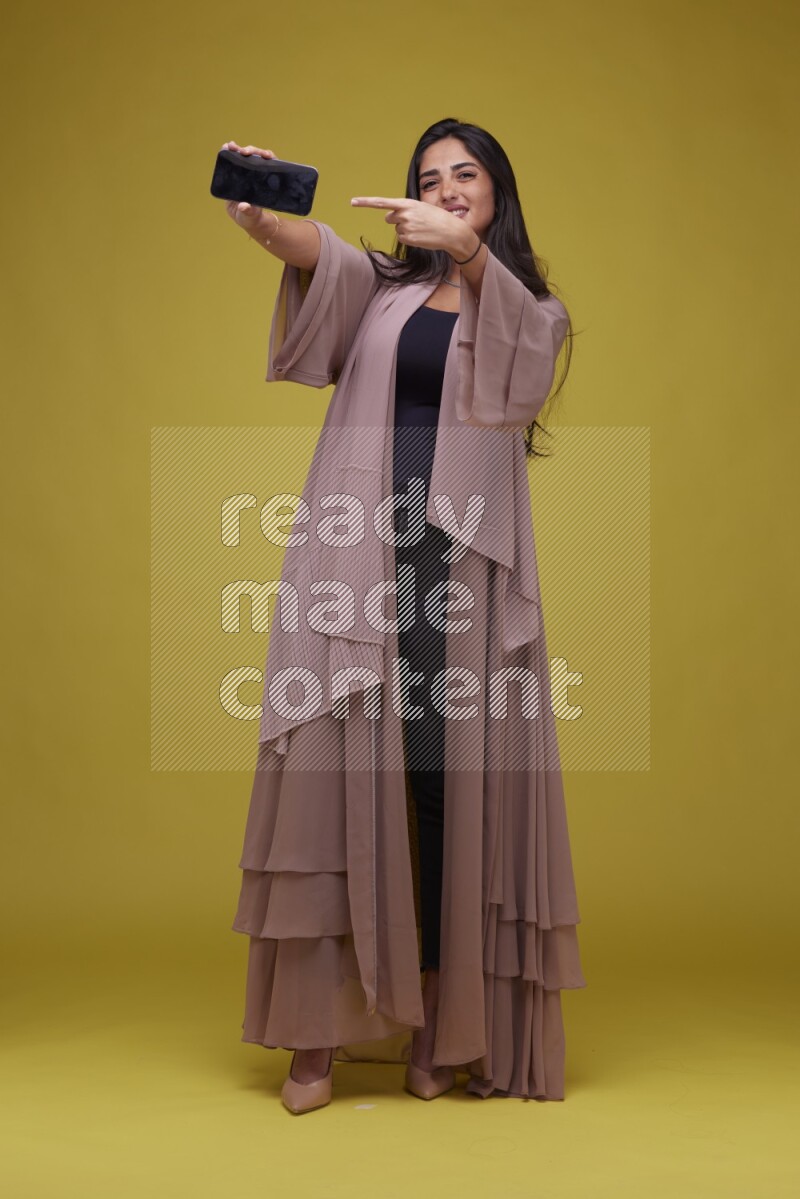 A woman Showing SmartPhone Screen on a Yellow Background wearing Brown Abaya