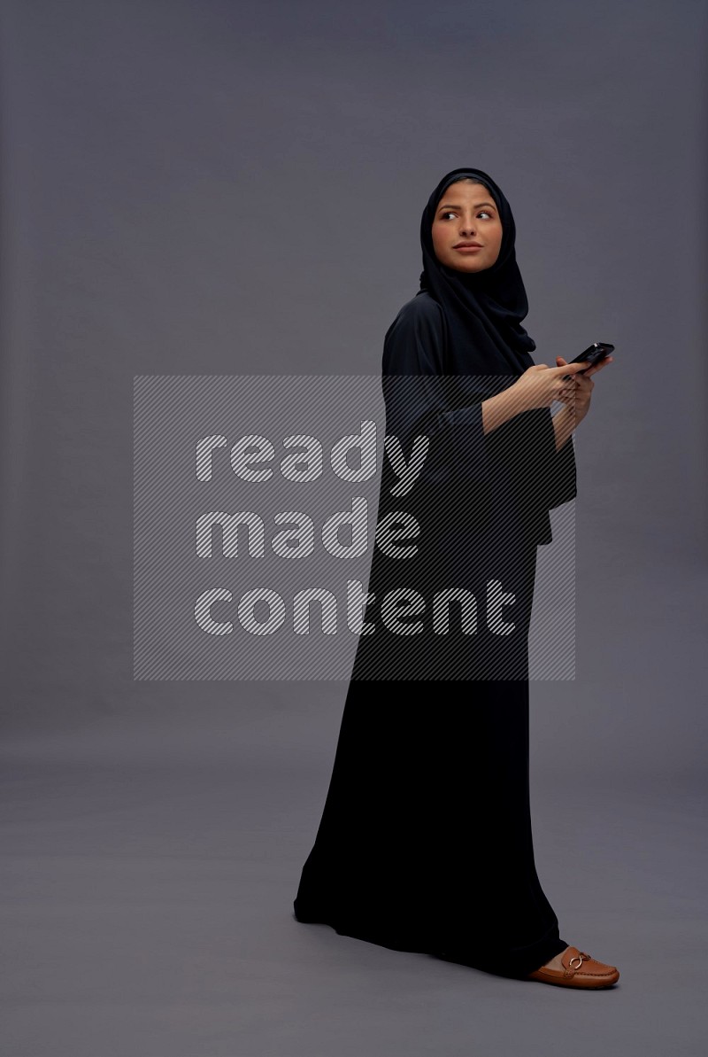 Saudi woman wearing Abaya standing texting on phone on gray background