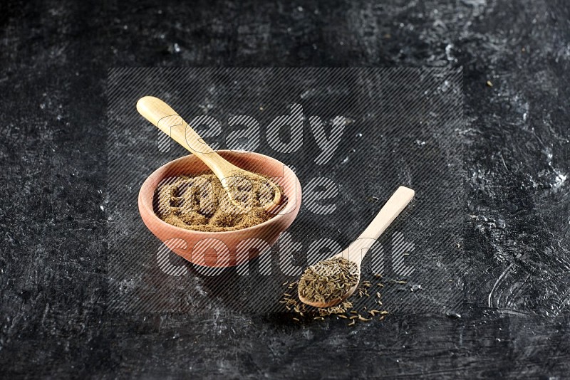 A wooden bowl and 2 wooden spoons full of cumin powder and cumin seeds on a textured black flooring