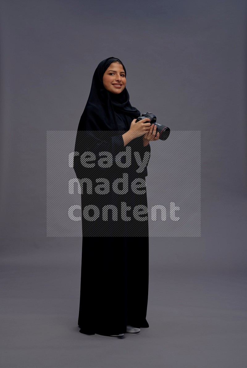 Saudi woman wearing Abaya standing holding Camera on gray background