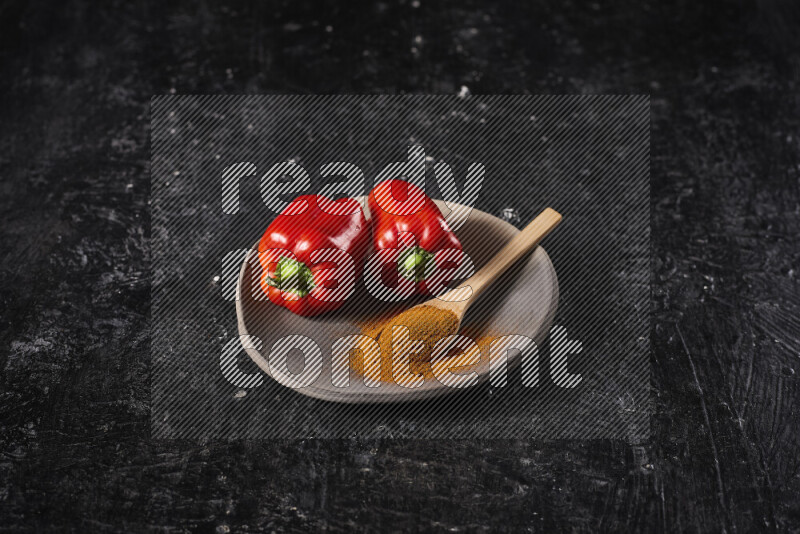 A wooden spoon full of ground paprika powder with red bell peppers, all on a pottery plate on black background