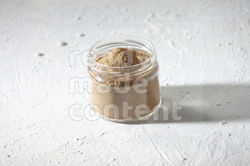 A glass jar full of cumin powder on textured white flooring