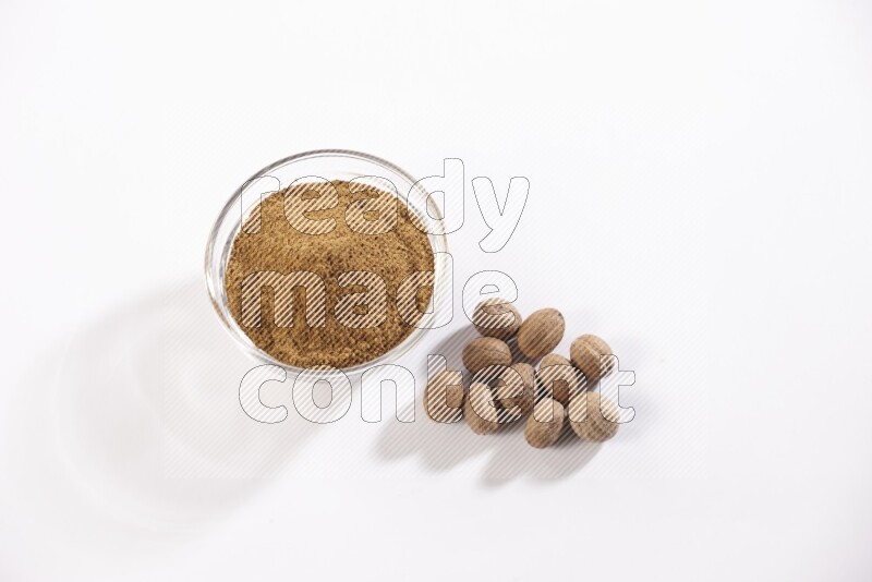 A glass bowl full nutmeg powder with the whole seeds beside it on a white flooring