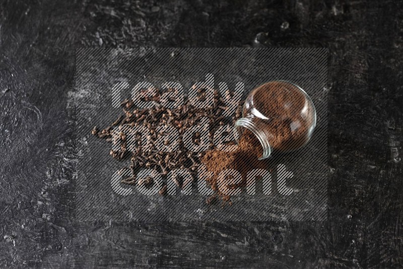 A flipped glass spice jar full of cloves powder and powder came out of it with cloves spread on textured black flooring