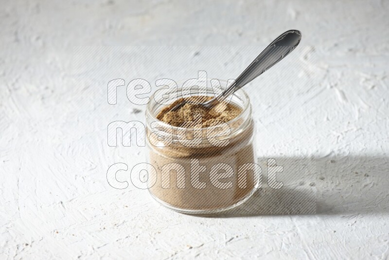 A glass jar and a metal spoon full of cumin powder on textured white flooring