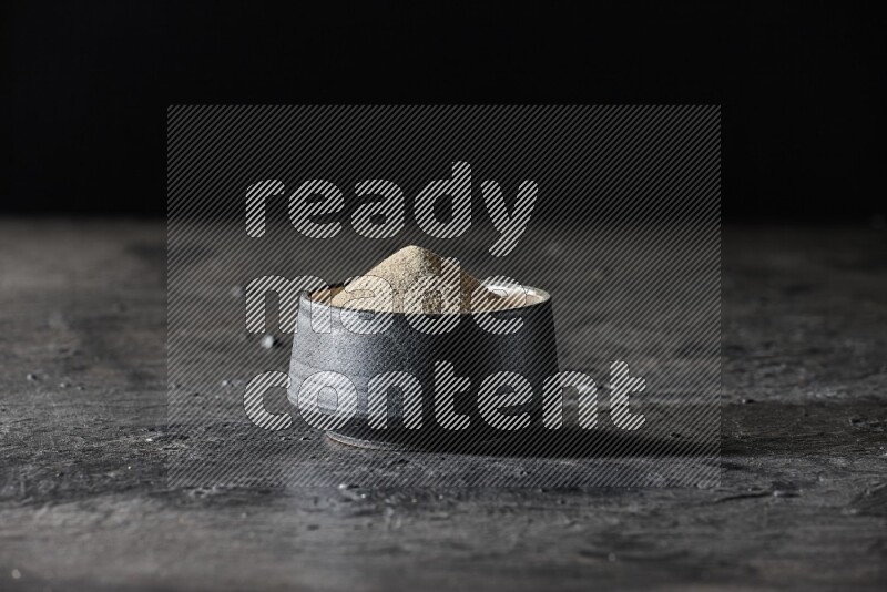 Black pottery bowl full of white pepper powder on textured black flooring