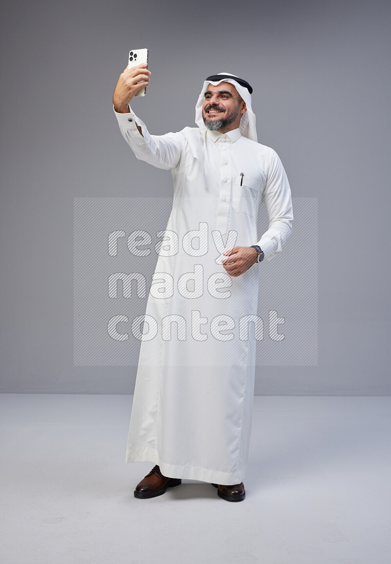 Saudi man Wearing Thob and white Shomag standing taking selfie on Gray background