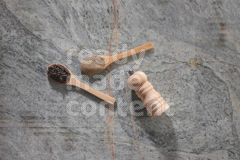2 wooden spoons, one full of black pepper powder and the other full of black pepper beads and wooden grinder on a marble flooring