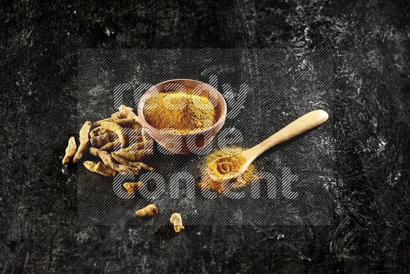 A wooden bowl and a wooden spoon full of turmeric powder with dried turmeric fingers on textured black flooring