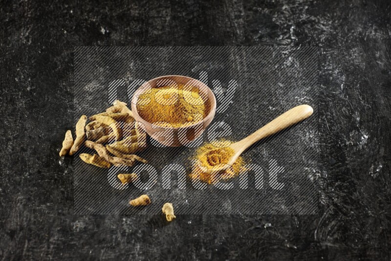 A wooden bowl and a wooden spoon full of turmeric powder with dried turmeric fingers on textured black flooring