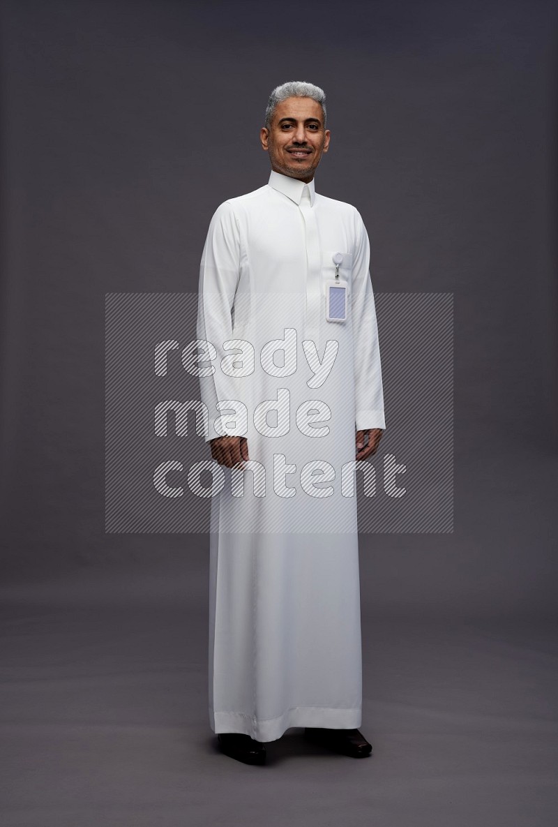 Saudi man wearing thob with pocket employee badge standing interacting with the camera on gray background