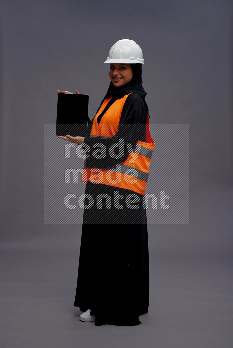 Saudi woman wearing Abaya with engineer vest standing showing tablet to camera on gray background