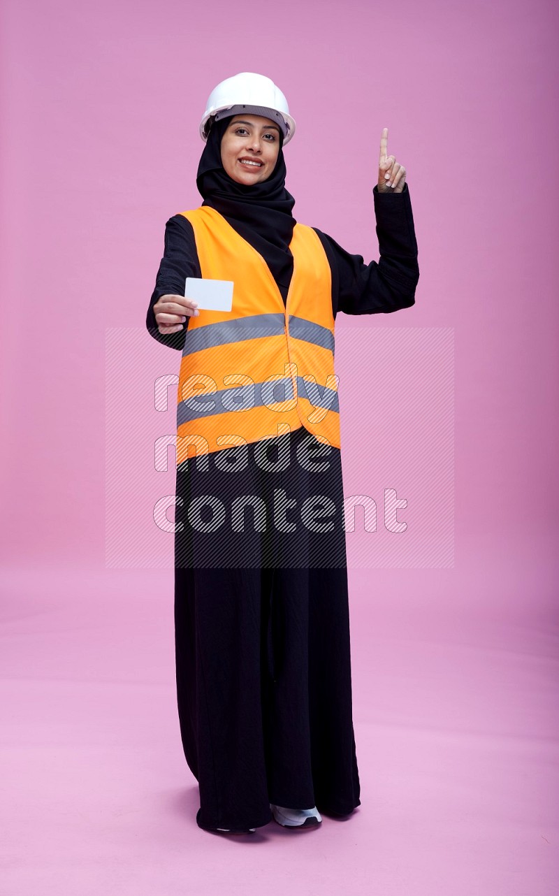 Saudi woman wearing Abaya with engineer vest and helmet standing holding ATM card on pink background