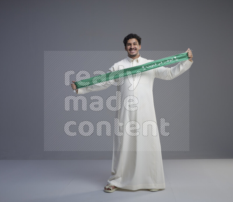 A Saudi man standing wearing thob holding Saudi flag scarf on gray background