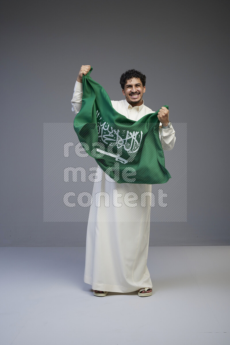 A saudi man standing wearing thob holding big saudi flag on gray background