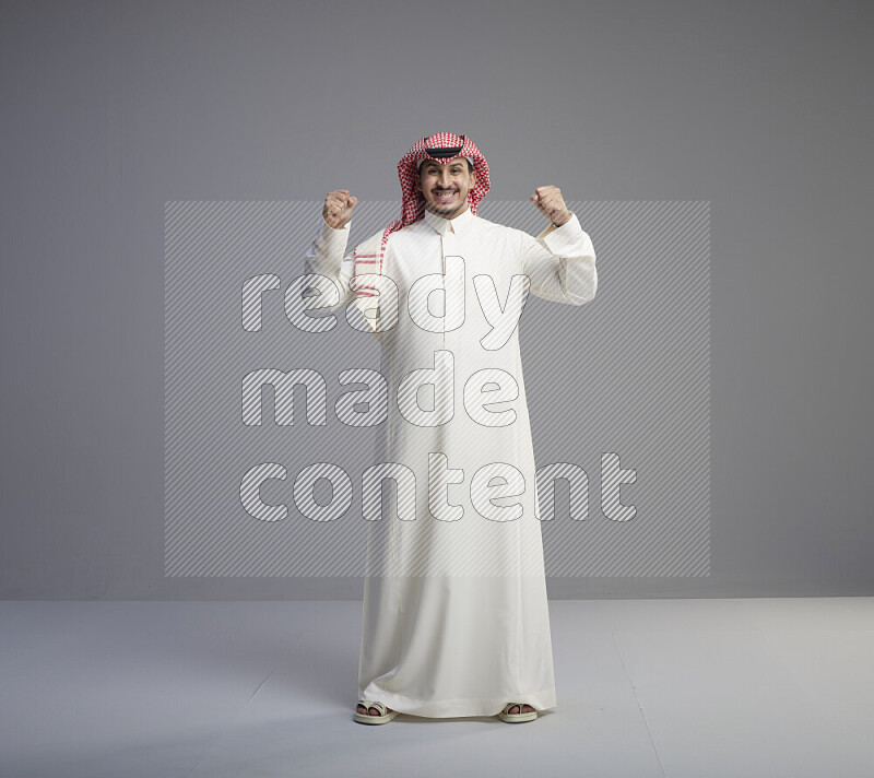 A Saudi man standing wearing thob and red shomag interacting with the camera on gray background