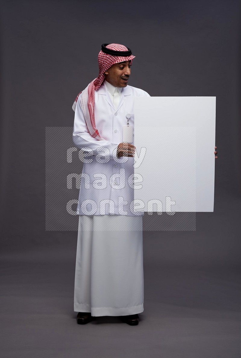 Saudi man wearing thob with lab coat and shomag with pocket employee badge standing holding board on gray background