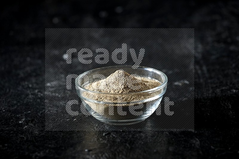 A glass bowl full of cardamom powder on textured black flooring