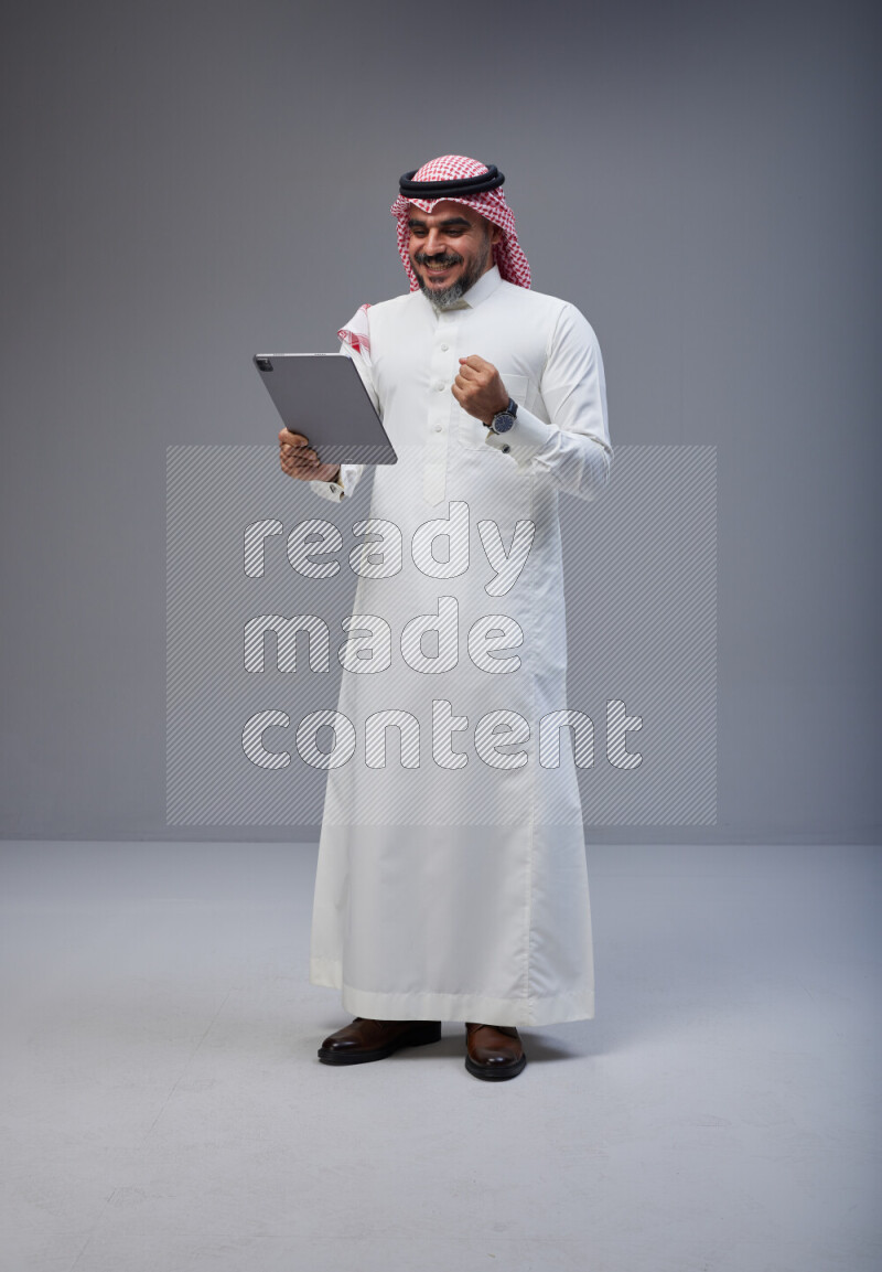 Saudi man Wearing Thob and red Shomag standing working on tablet on Gray background