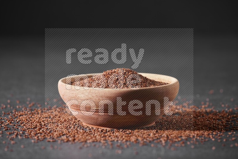 A wooden bowl full of garden cress seeds surrounded by seeds on a black flooring