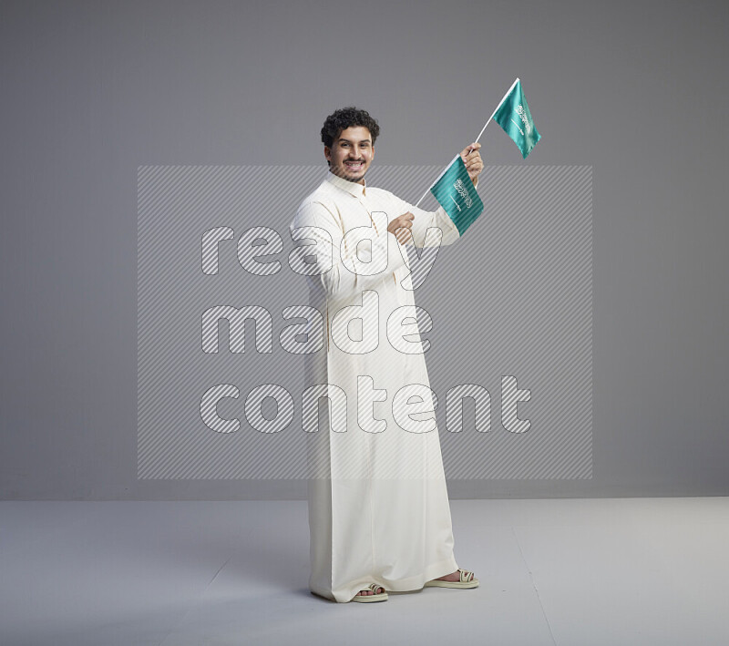 A Saudi man standing wearing thob holding small Saudi flag on gray background