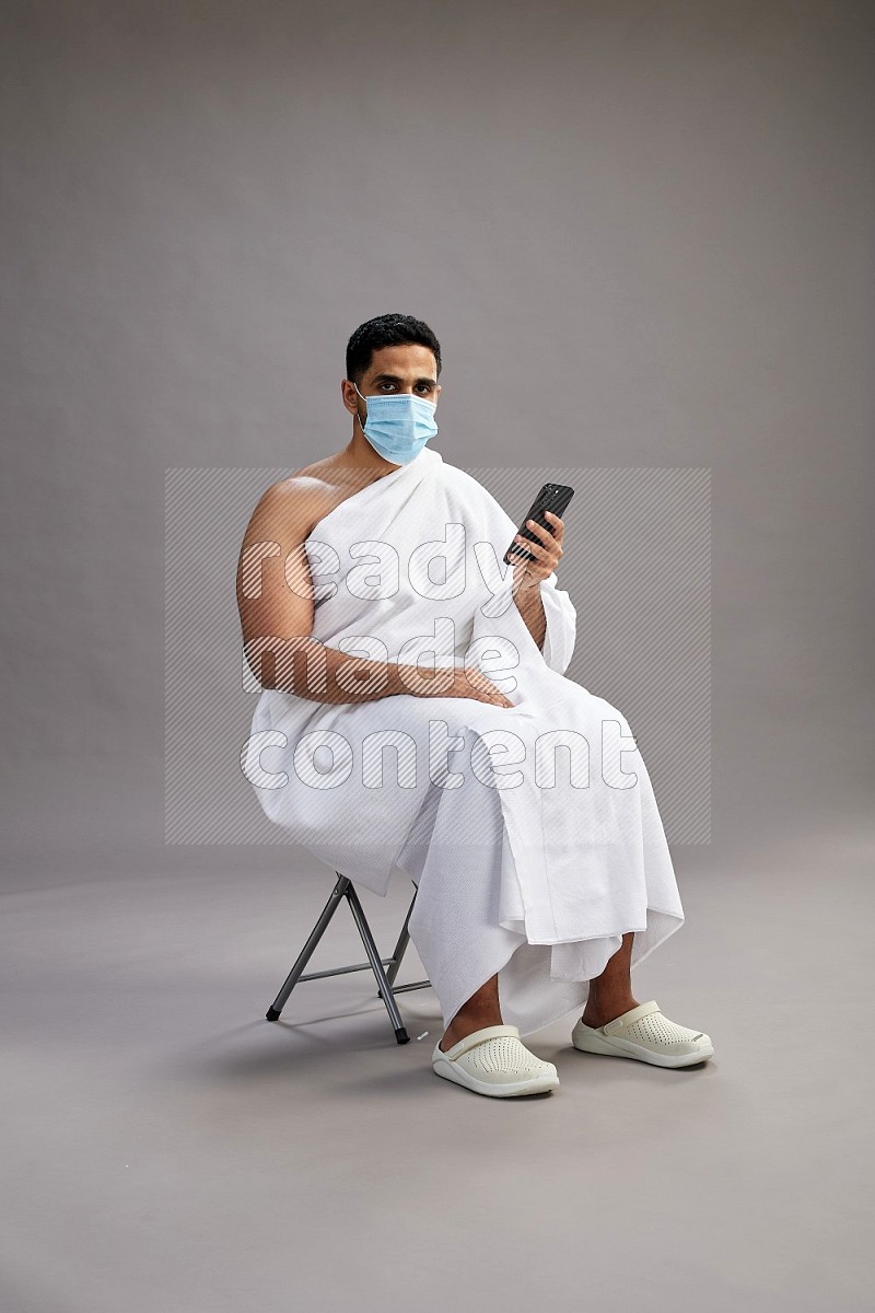 A man wearing Ehram with face mask sitting on chair texting on phone on gray background